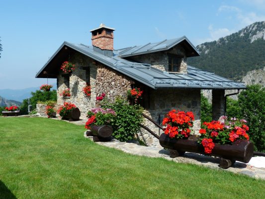 Foto von einem kleinen Ferienhaus in den Bergen mit roten Blumen und grüner Wiese davor.